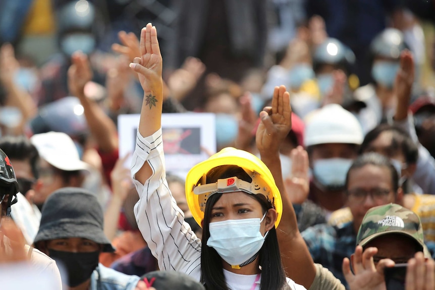 A young woman wearing a face mask and yellow helmet raises three fingers in a crowd on sunny day.