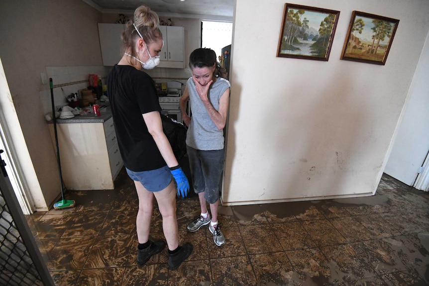 Two women stand in a house that has been muddied by floodwaters - one is wearing a mask, the other holds her face in anguish.