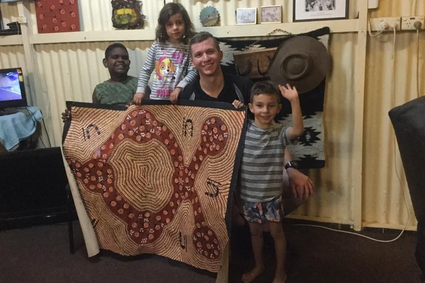 Three children holding an Indigenous painting stand with a young man, who is kneeling to their height.