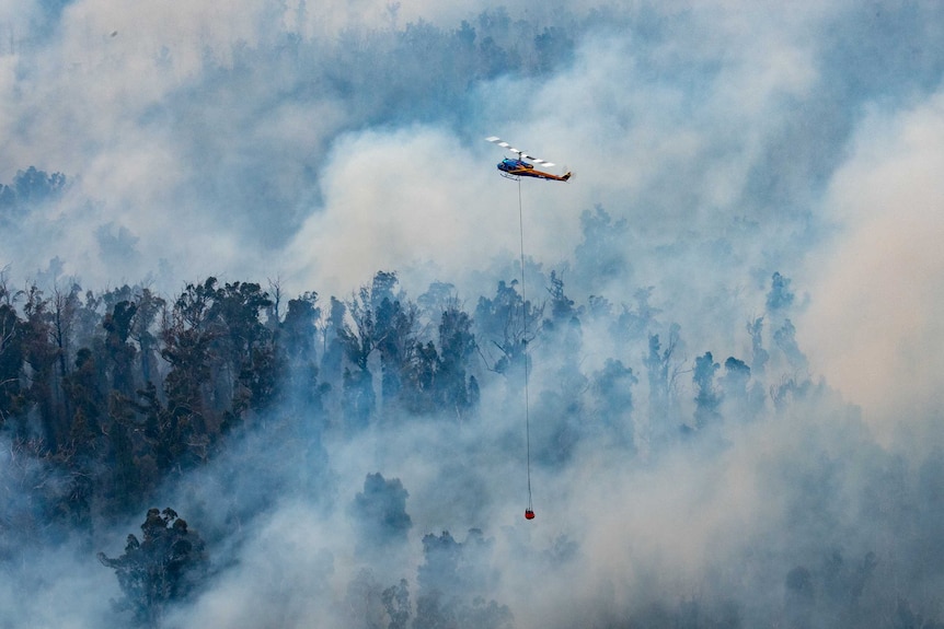 A helicopter flies through smoke over a burning forest.