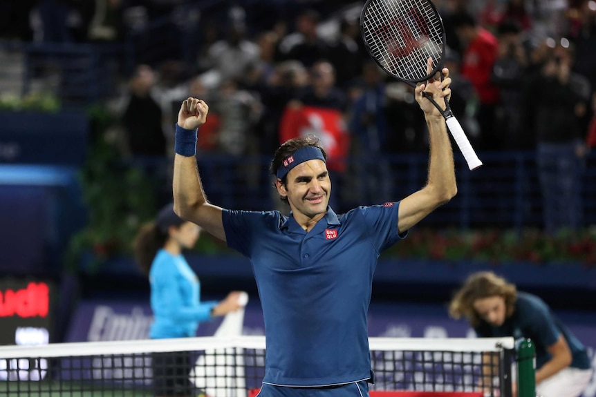 Tennis player raises his arms in triumph as he stands in front of the net at the end of a match.