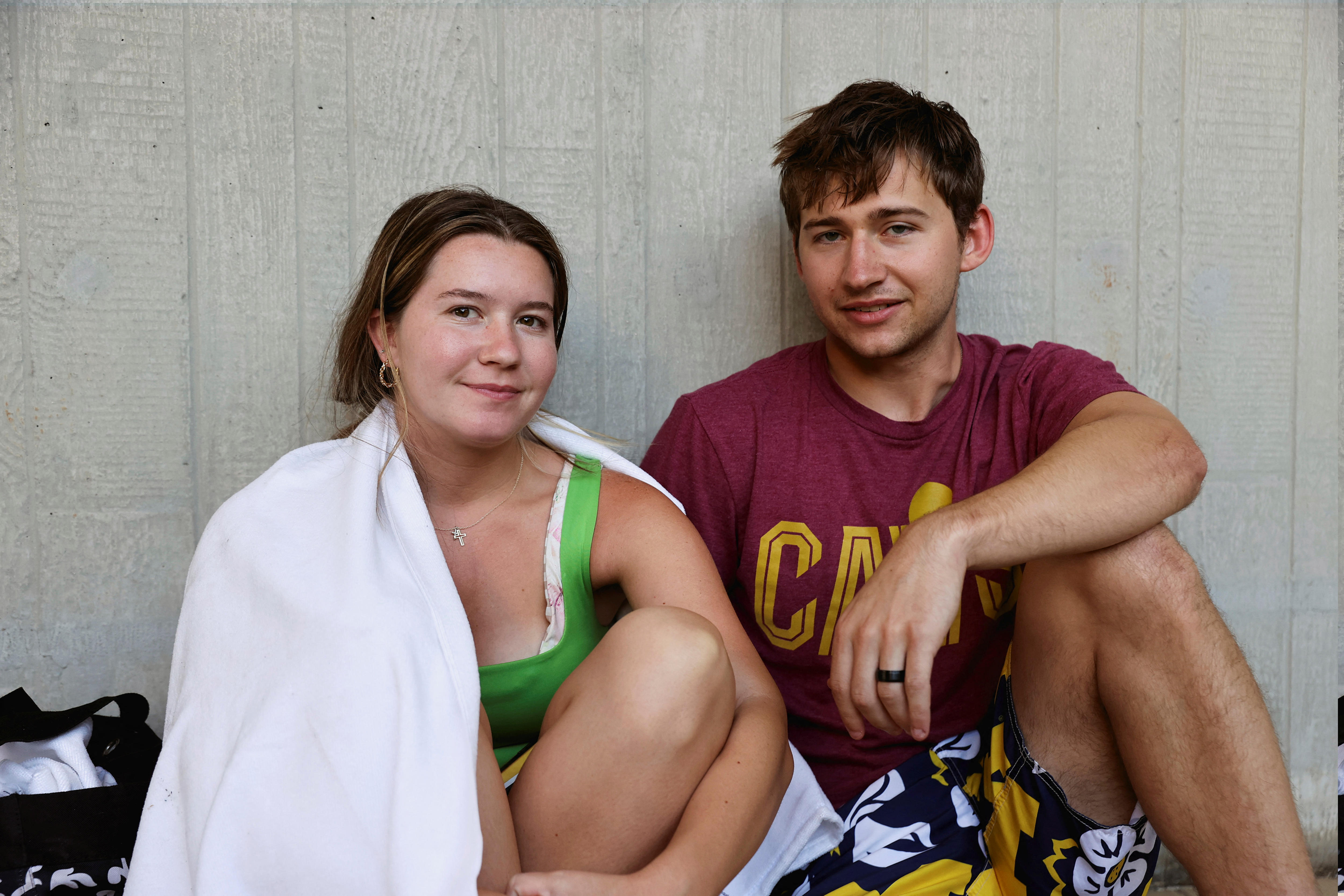 A young couple sit down at airport, woman in a green tank top covered in a blanket, a guy in red shirt, floral board shorts 