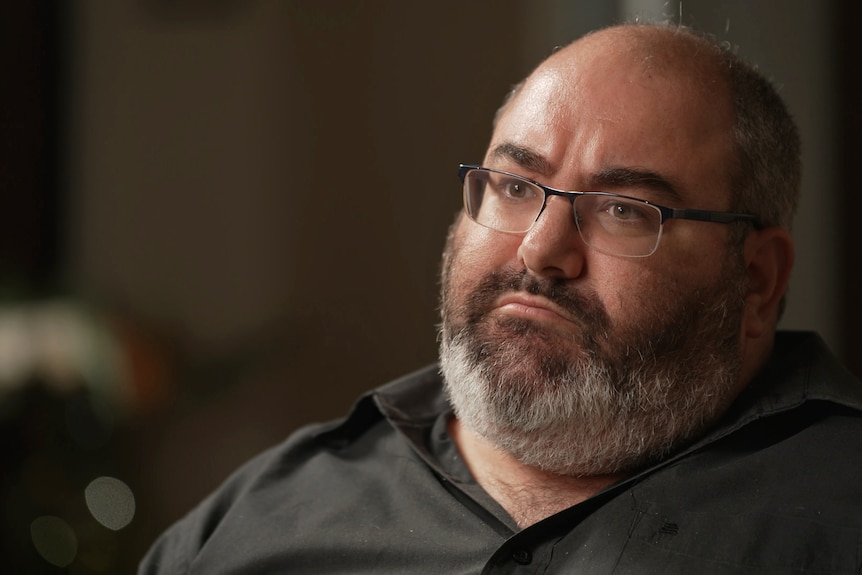 A man sitting indoors, looks to the side with a neutral expression.