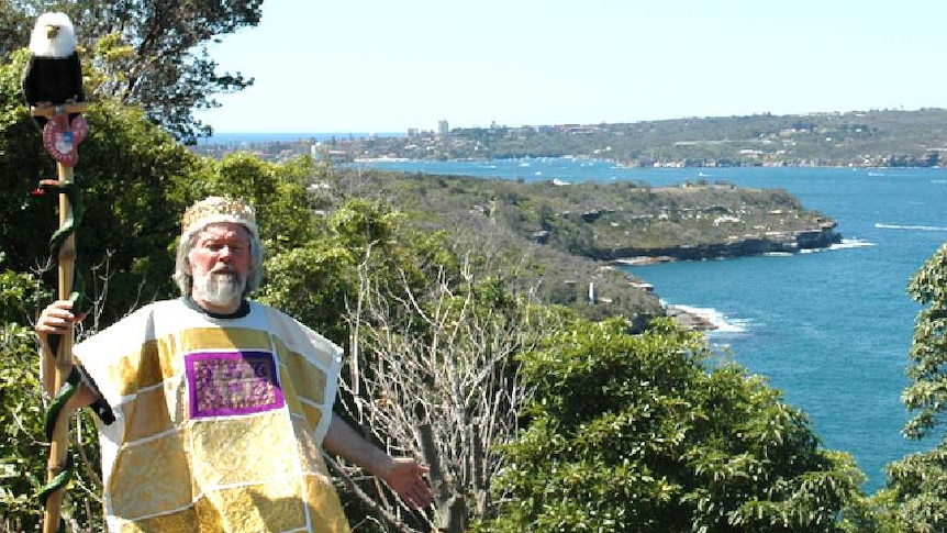 Paul Delprat stands in front of a coastline holding a staff and wearing a yellow outfit.