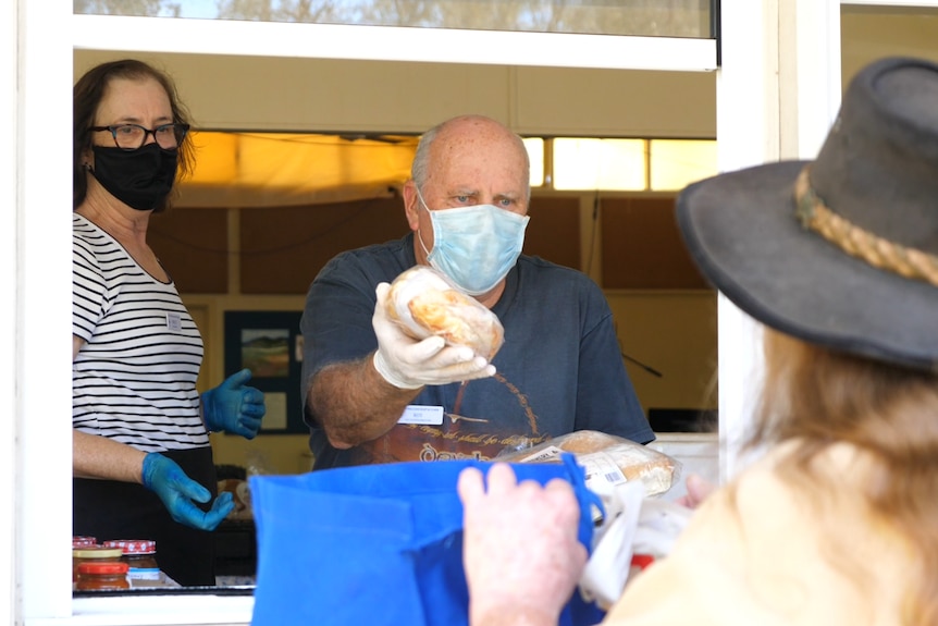An older man wearing a mask hands some wrapped bread rolls to a woman.