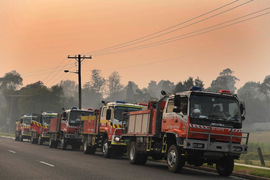 Five fire trucks parked on the side of a road, with smoke all around.