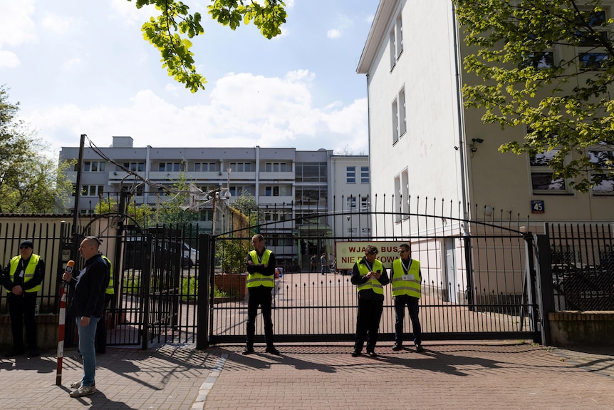 A small number of men in work clothes and yellow hi-vis vests stand in front of a spiked gate to a diplomatic building.