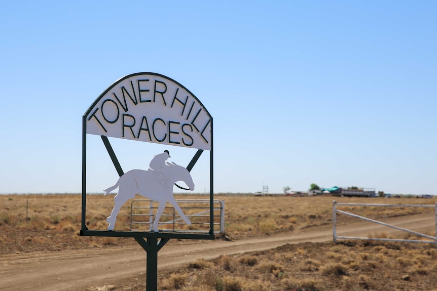 A metal sign at the entrance of the Tower Hill racecourse showing a horse and a rider, bare flat land behind, blue skies.