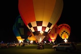 Huge brightly-coloured hot air balloons light up the sky. People gather around the baskets, some cars in foreground.