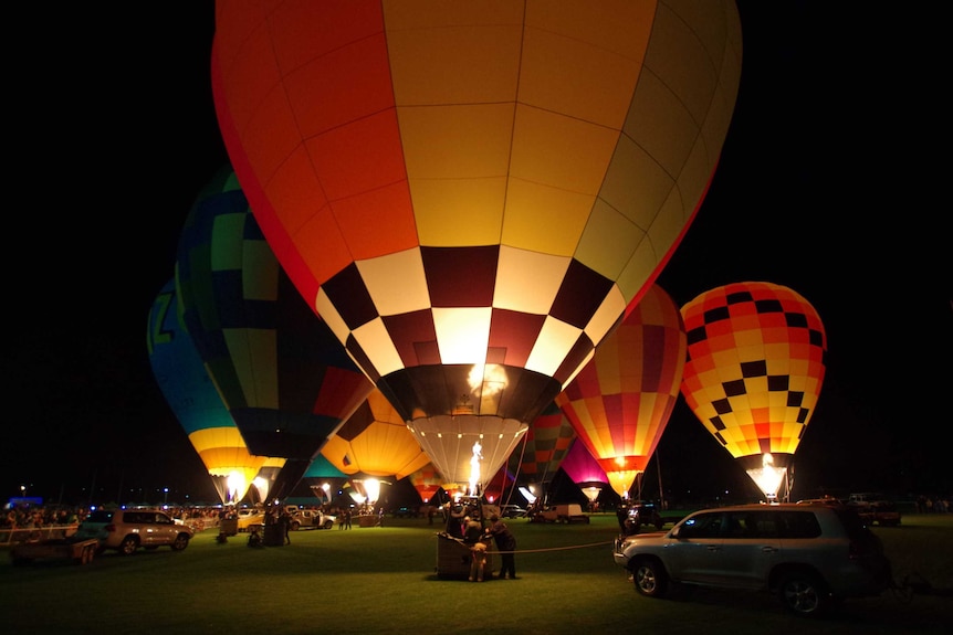 Huge brightly-coloured hot air balloons light up the sky. People gather around the baskets, some cars in foreground.