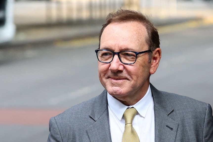Spacey, wearing a blue suit and pink tie, gives a closed lipped smile