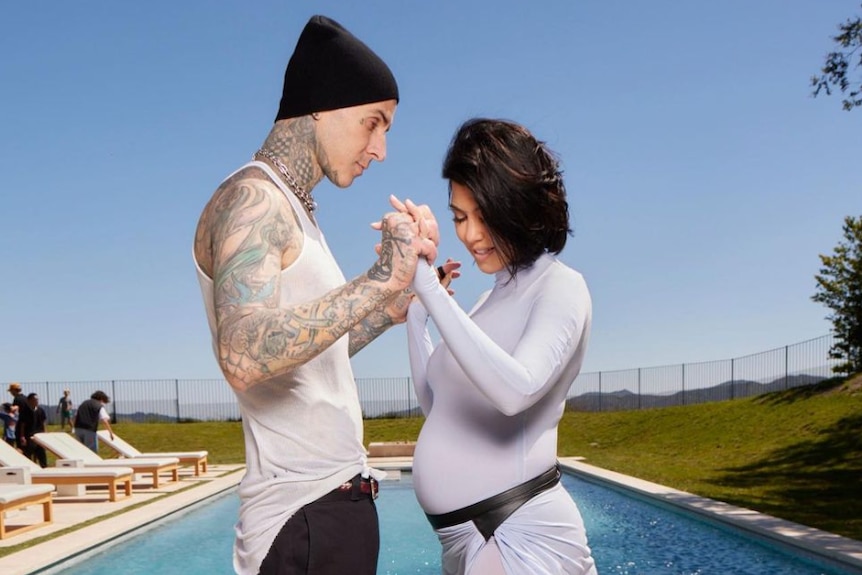 Travis Barker and a pregnant Kourtney Kardashian Barker hold hands in front of a pool.