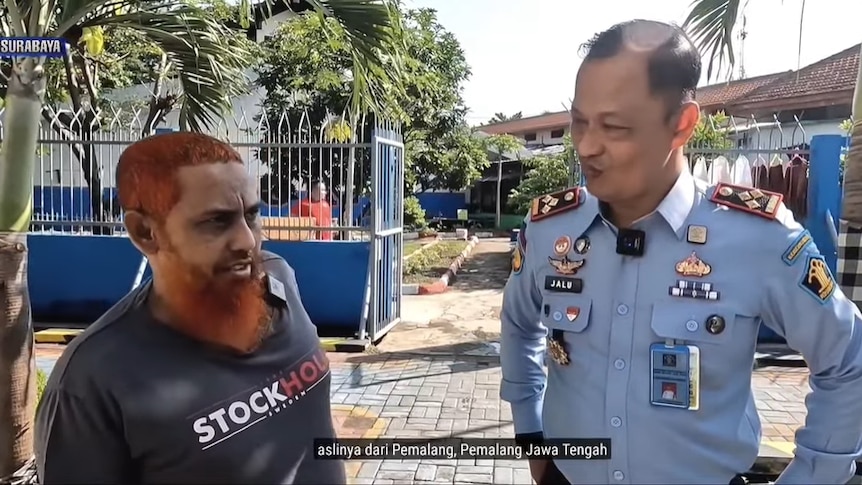 Umar Patek looks on while being interviewed in prison by a staff member surrounded by palm strees and pathways