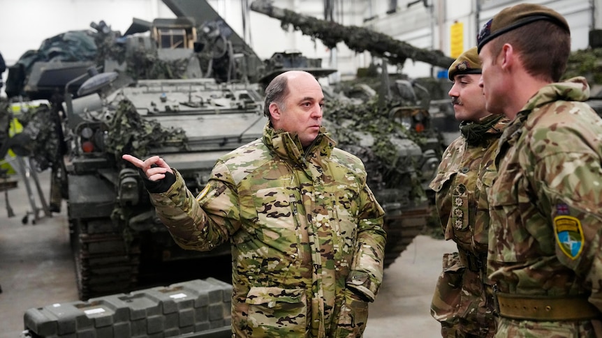 A partly bald man with greying hair in green military camoflauge gestures in front of tanks