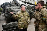 A partly bald man with greying hair in green military camoflauge gestures in front of tanks