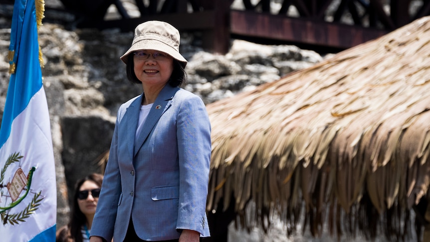 Tsai Ing-Wen, wearing a blue linen blazer and beige bucket hat, smiles next to a flag and an outdoor structure