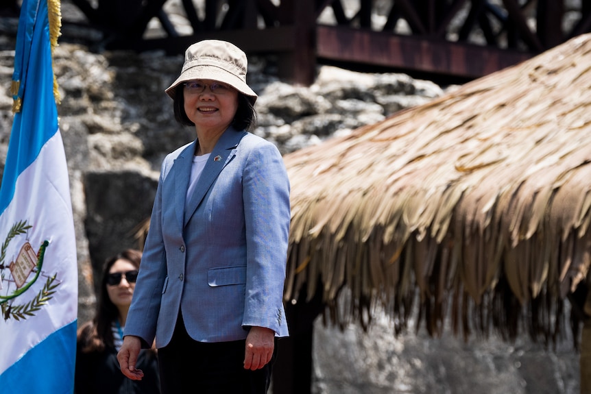 Tsai Ing-Wen, wearing a blue linen blazer and beige bucket hat, smiles next to a flag and an outdoor structure