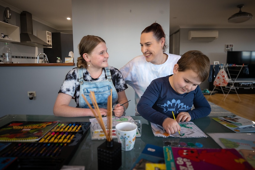 A young woman sits at a table with her two primary school children who are painting