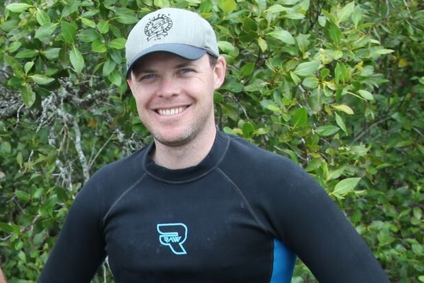 Dr Ben Gilby stands in Noosa River with bush behind him wearing rash vest with hands on hips