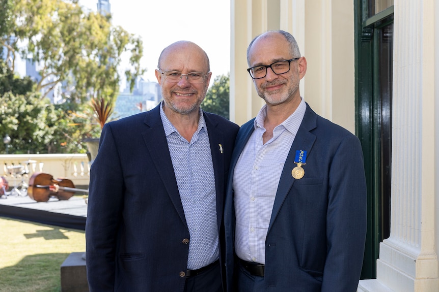 Michael Barnett stands with his partner Gregory. They both wear dark suits, blue shirts and glasses.