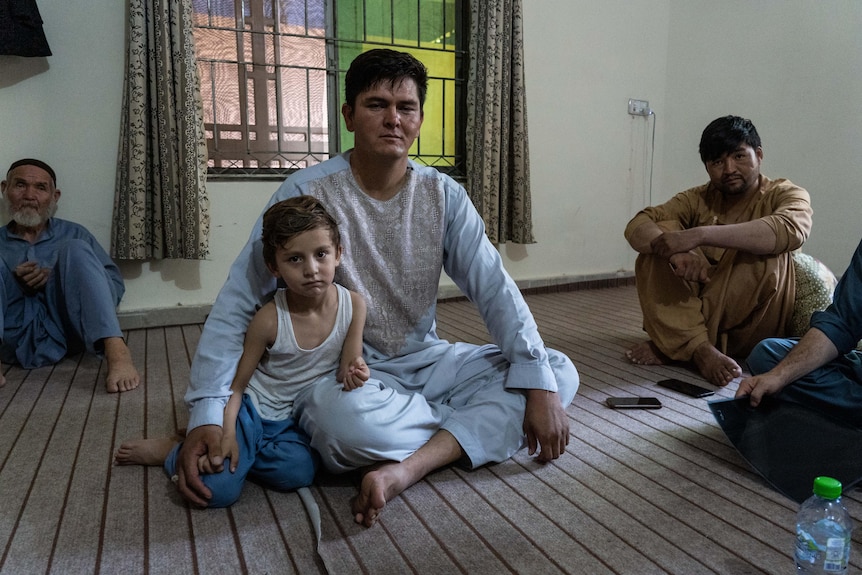 A young boy sits with his father, who is cross-legged on the ground. 