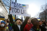 A woman at a protest holds a sign which reads 'Help before it's too late', and has a Ukrainian flag on it.