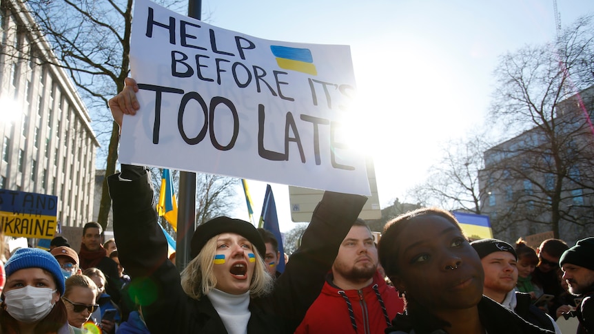 A woman at a protest holds a sign which reads 'Help before it's too late', and has a Ukrainian flag on it.