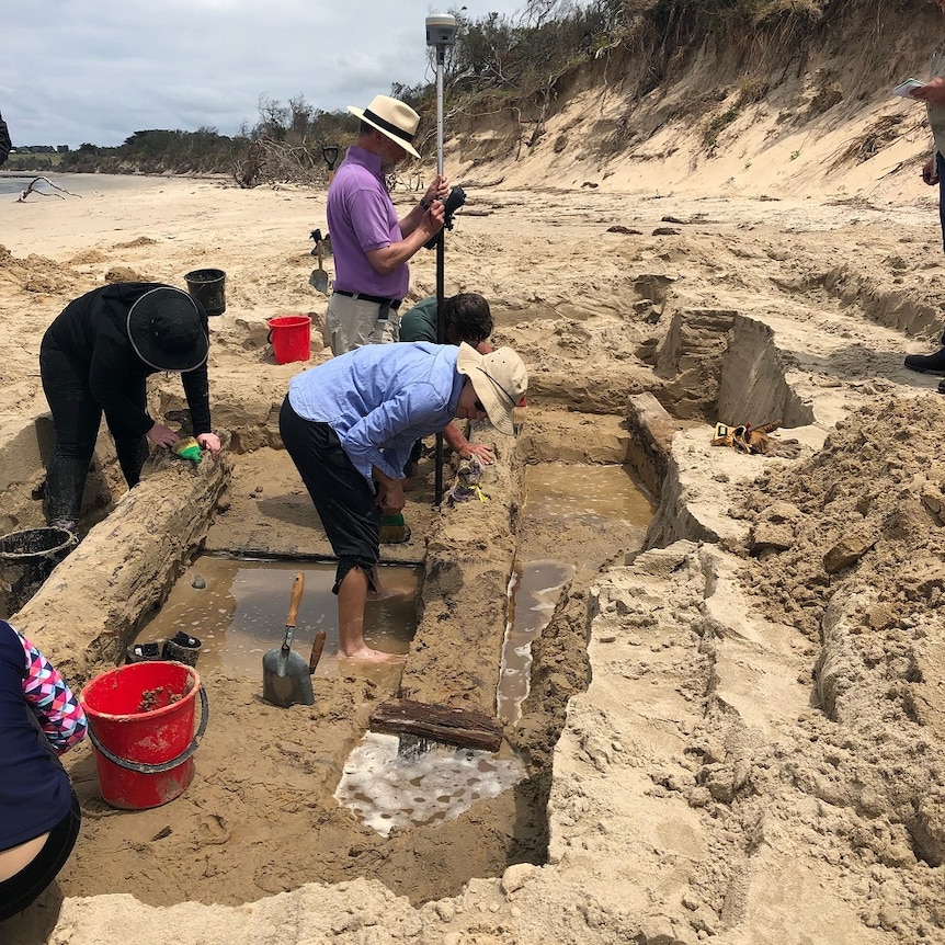 A group of workers stand in the sand digging around pieces of wood from a 19th century shipwreck.