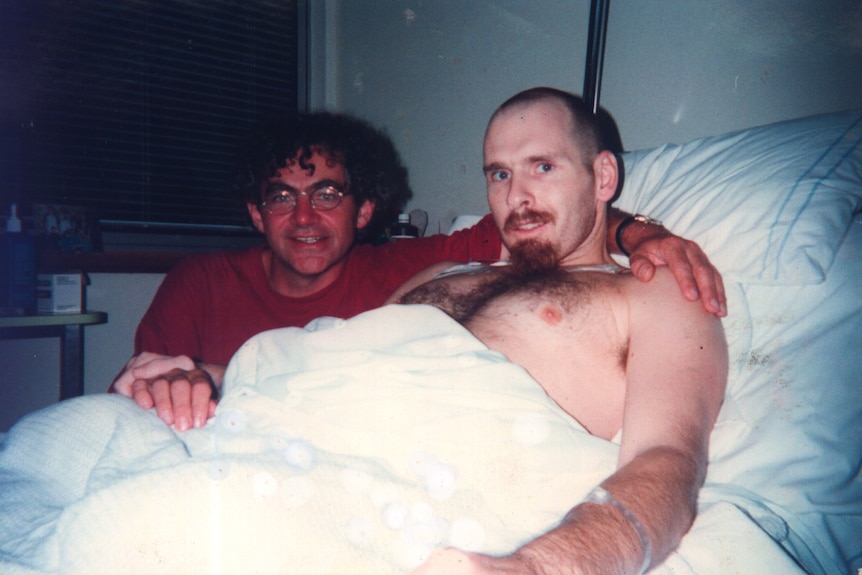 One man in a hospital bed, another with his arm around his friend.