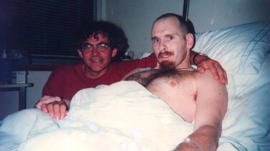 One man in a hospital bed, another with his arm around his friend.