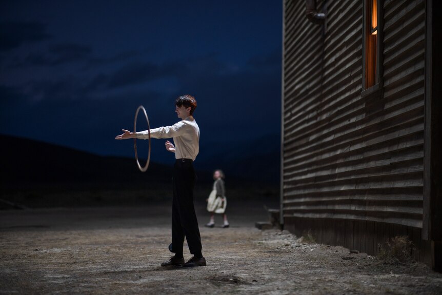 An effete teenage boy in a white collared shirt stands outside balancing a hula hoop on his outstretched arm