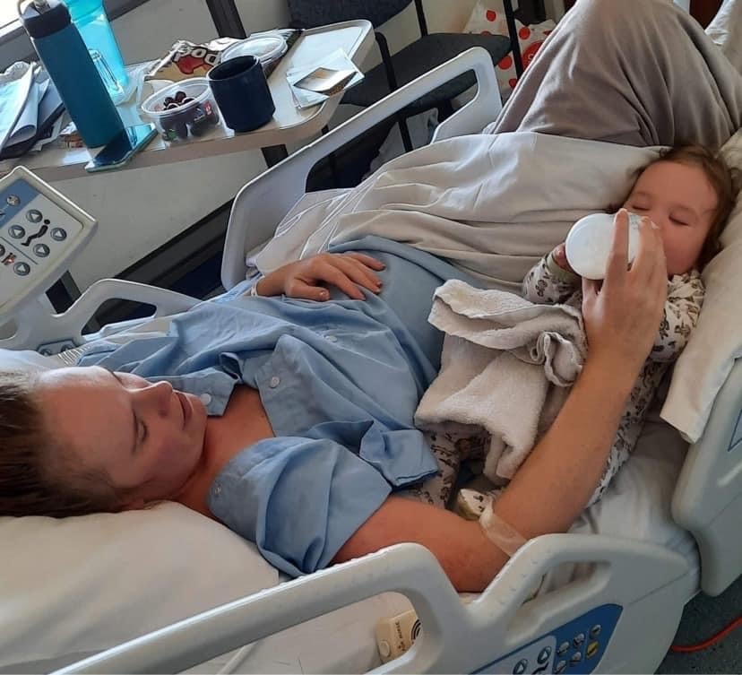 A mum laying in a hospital bed in blue hospital robes feeding a bottle of milk to a baby