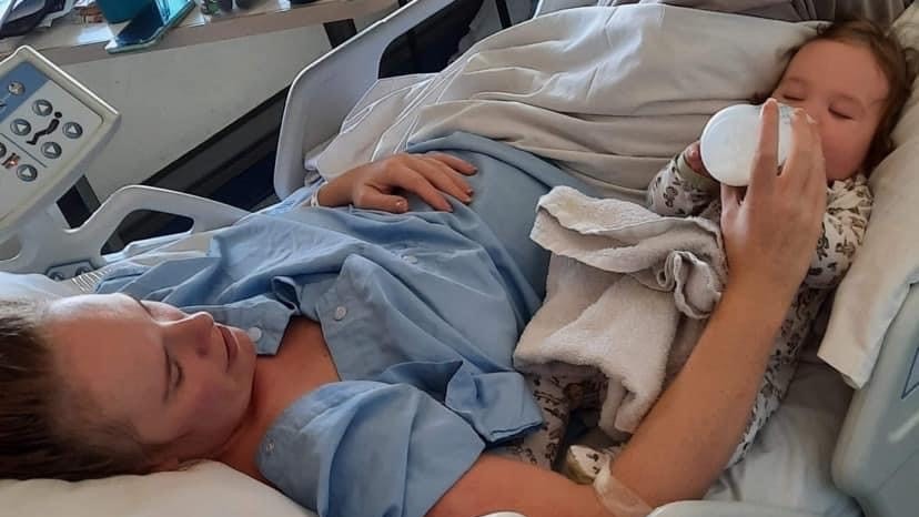 A mum laying in a hospital bed in blue hospital robes feeding a bottle of milk to a baby