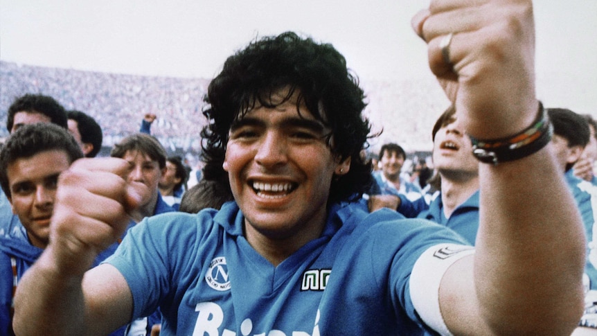 Maradona in his sky blue jersey and his curly hair falling almost to his shoulders grins at the camera and raises both fists.