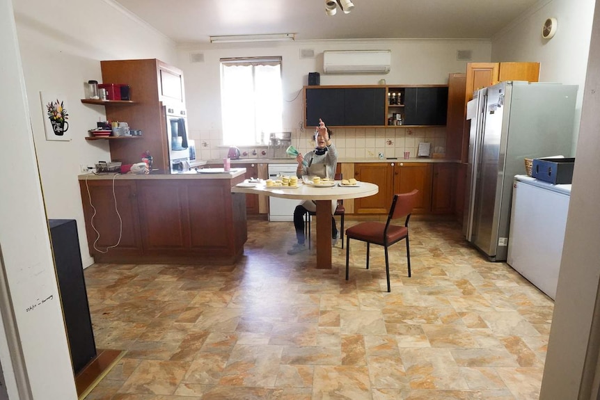 Camila De Ville sits at a small kitchen table assembline alfajores in a large old kitchen with vinyl floor and wooden cabinets.