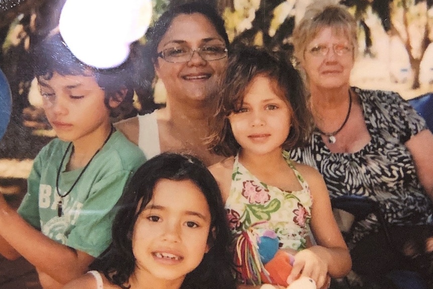 Image is a picture of a polaroid with Amishah on her mother's lap, centre, with her grandmother right, brother left and cousin.