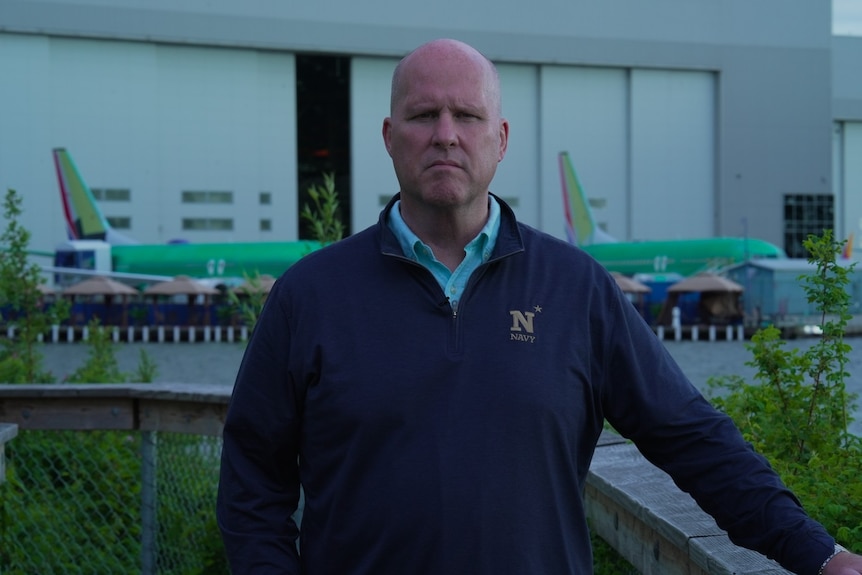A bald man in a jumper in front of an aeroplane factory.