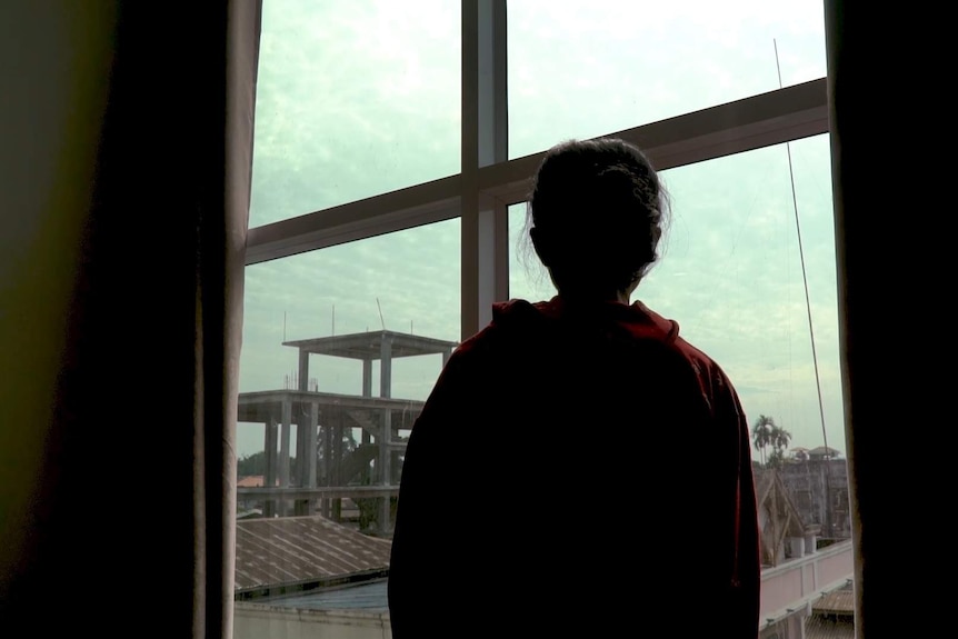 A trafficking victim from Myanmar stands facing a window. The sky is overcast and you can see rusted roofs.