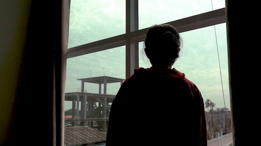 A trafficking victim from Myanmar stands facing a window. The sky is overcast and you can see rusted roofs.