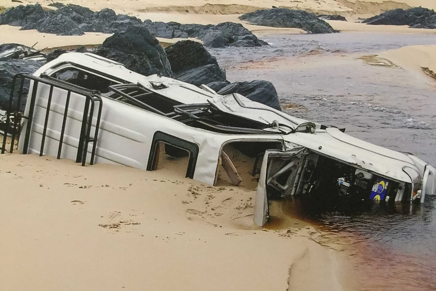 A ute on its side lying in water and sand near a beach, surrounded by rocks, with bonnet open