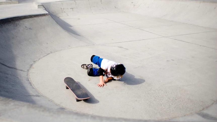 A young child on the ground after falling off their skateboard at a skatepark