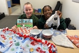 Two smiling girls hold materials while sitting at a table filled with blankets and bowls