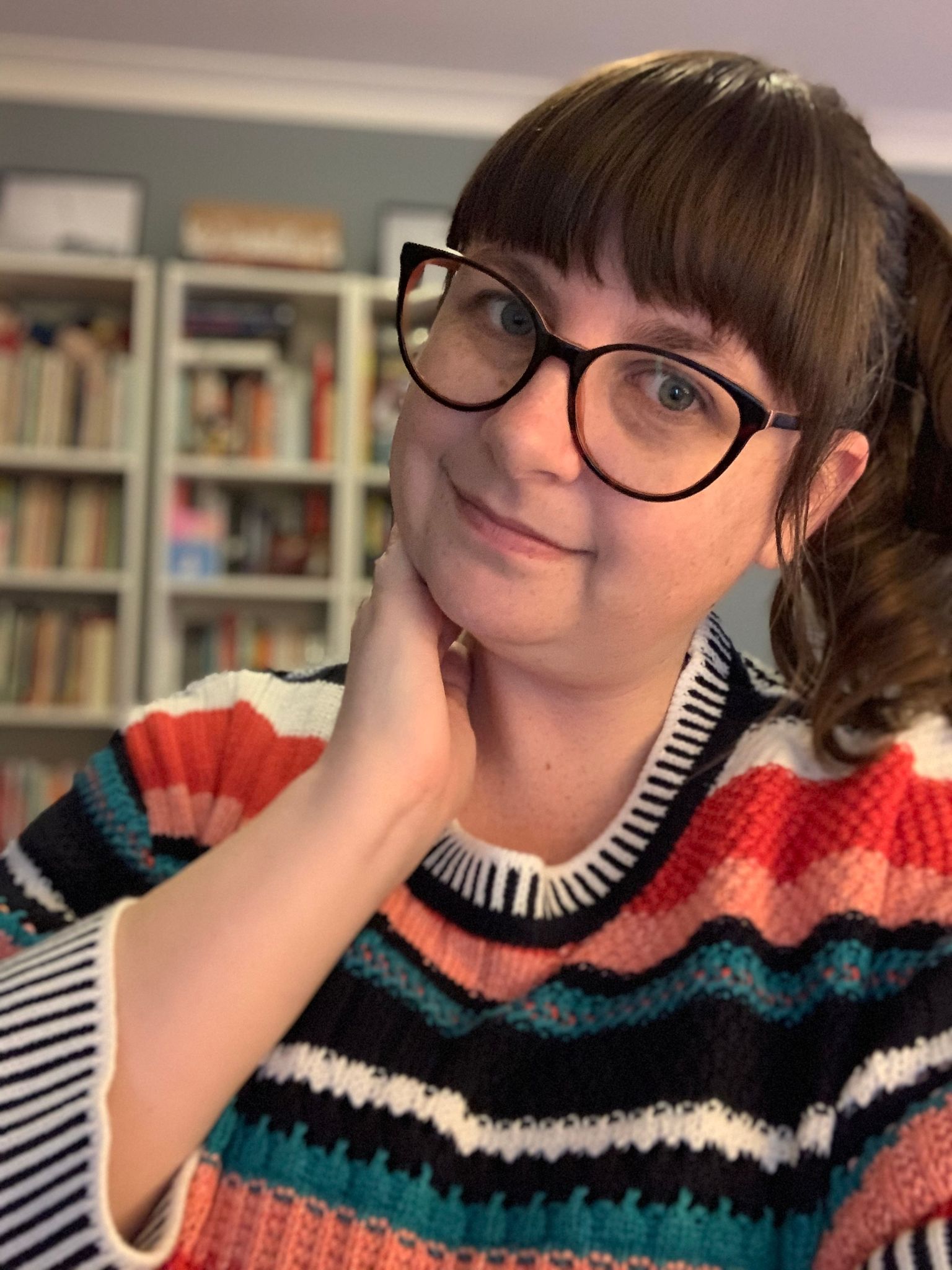 A brown-haired woman in her late 30s with a fringe and ponytail and glasses looks into the camera, a hand under her chin