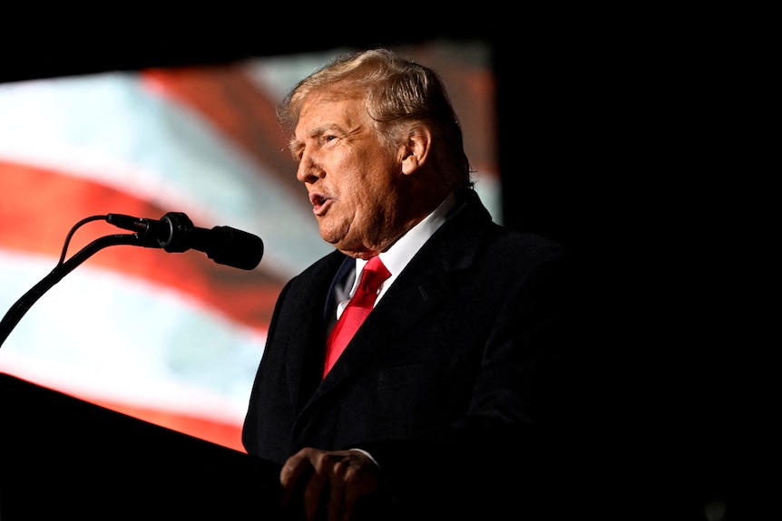 Donald Trump speaks into a microphone in front of a projected american flag visual