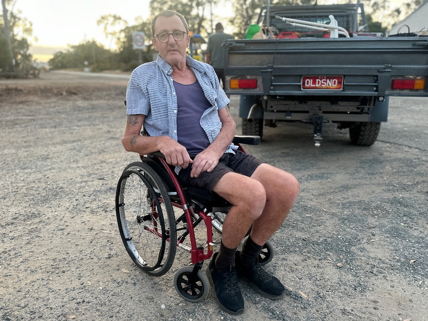 An older man sits in a wheelchair on a gravel road.