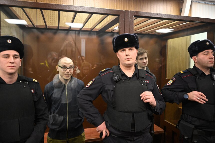 Three male guards stand in front of a glass room inside a courtroom, in which two men stand and smile slightly