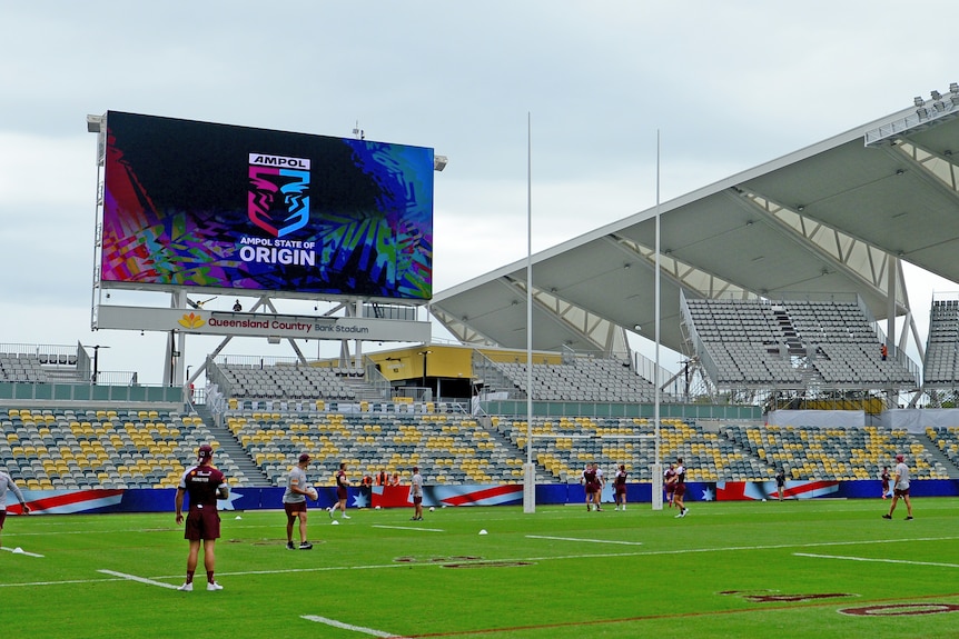 A group of Queensland players run around an empty stadium with a big State of Origin sign on the scoreboard behind them.