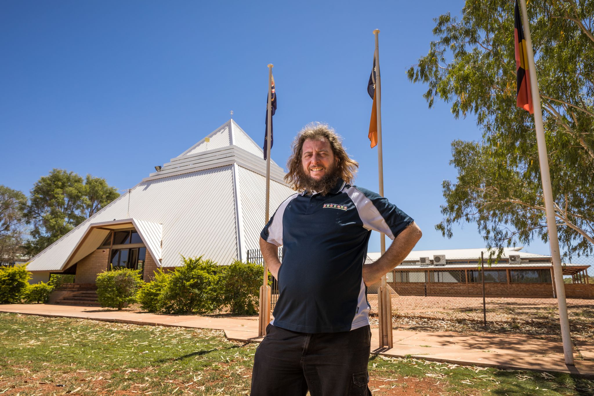 A man with long hair in a polo shirt stands in front of a pyramid shaped building and flag poles.