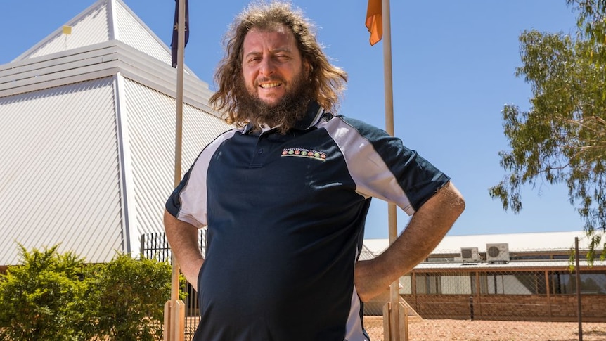 A man with long hair, wearing a polo shirt, stands in front of a pyramid-shaped building and flag poles.