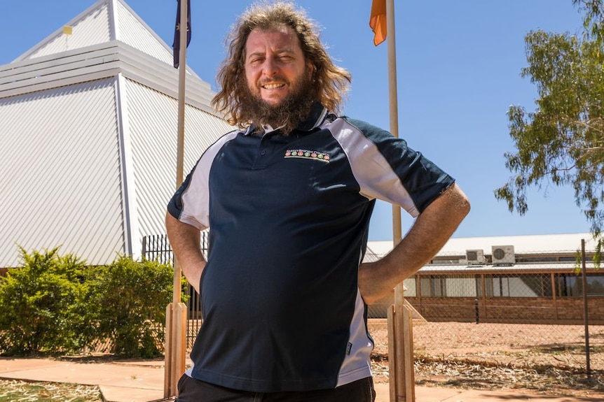 A man with long hair in a polo shirt stands in front of a pyramid shaped building and flag poles.